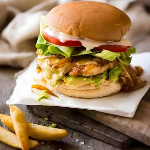 A chicken burger with avocado, caramelized onions, melted cheese, lettuce, tomato, and mayo, with fries.